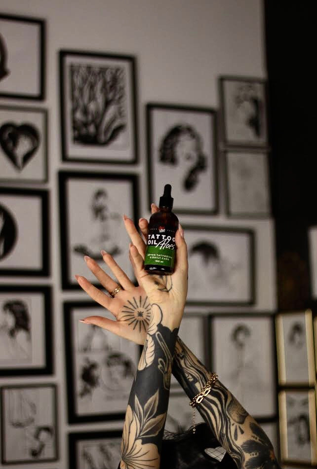 Kobiece dłonie na tle galerii czarno-bialych rysunków w ramkach trzymają Loveink Tattoo Oil Aloes 