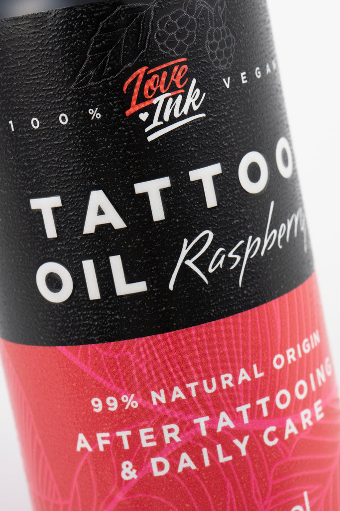 Zbliżenie na etykietę butelki oleju do tatuaży o zapachu malinowym od marki Love Ink. Na czarnej etykiecie znajduje się biały napis "Tattoo Oil Raspberry" oraz czerwone logo Love Ink. W dolnej części różowej etykiety podano, że produkt jest w 99% pochodzenia naturalnego i przeznaczony do pielęgnacji po tatuażu oraz do codziennego użytku. Design etykiety zawiera delikatny wzór malin i liści, co podkreśla naturalne składniki produktu.